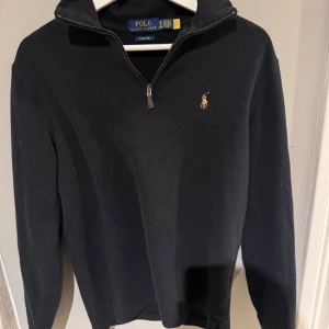 Svart tröja med dragkedja från Ralph Lauren - Säljer en stilren svart tröja från Ralph Lauren med dragkedja och klassisk logga på bröstet. Perfekt för en avslappnad men snygg look. Tröjan har långa ärmar och en bekväm passform.