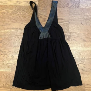 Svart klänning med läderdetaljer - Snygg svart klänning med breda axelband i läderimitation. Den har en djup v-ringning och en lös passform som ger en avslappnad look. Perfekt för en kväll ute eller en speciell tillställning.