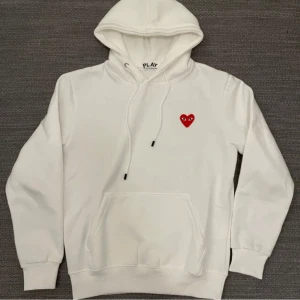 Vit hoodie med rött hjärta från Comme des Garçons - Säljer en stilren vit hoodie från Comme des Garçons med ett ikoniskt rött hjärta på bröstet. Hoodien har en klassisk passform med långa ärmar och en praktisk magficka. Perfekt för en avslappnad look.