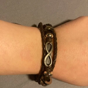 Brunt flätat armband med infinity-symbol - Snyggt brunt flätat armband med en silverfärgad infinity-symbol i mitten. Armbandet har flera lager och små silverfärgade detaljer som ger det en unik look. 