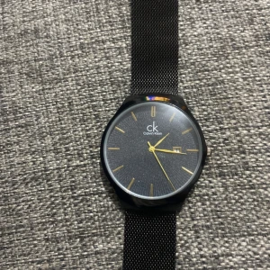 Svart klocka från Calvin Klein - Elegant svart klocka från Calvin Klein med en stilren urtavla och guldiga visare. Klockan har ett diskret datumfönster och ett svart mesh-armband som ger en modern look. Perfekt för den som gillar minimalistisk design.