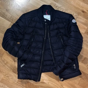 Svart pufferjacka från Moncler - Snygg svart pufferjacka från Moncler med dragkedja och quiltad design. Jackan har långa ärmar och en stilren look med Moncler-logga på ärmen. Perfekt för kyligare dagar.