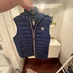 Moncler väst - Snygg blå dunväst från Moncler med dragkedja och huva. Perfekt för kyligare dagar. Västen har Monclers logotyp på bröstet och en stilren design.