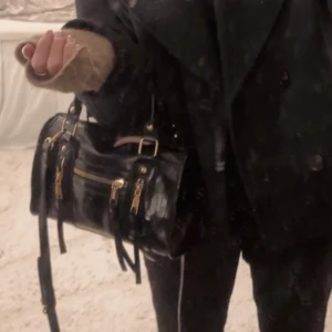 Svart axelväska med dragkedja - Snygg svart axelväska i skinn med guldfärgade detaljer och dragkedja framtill. Perfekt för att bära dina nödvändigheter med stil. Väskan har en praktisk storlek och justerbar axelrem.