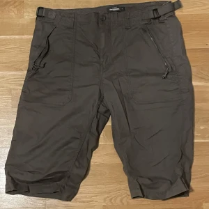 Bruna shorts från RevolutionRace - Snygga bruna shorts från RevolutionRace med praktiska fickor och justerbara remmar i midjan. Perfekta för utomhusaktiviteter med en bekväm passform och dragkedjor på fickorna. Passar bra för sommarens äventyr.