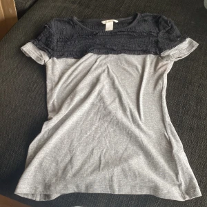 Grå och svart topp med spetsdetaljer - Säljer en snygg grå topp med svarta spetsdetaljer upptill. Toppen har korta ärmar och en bekväm passform. Perfekt för både vardag och fest! 🖤