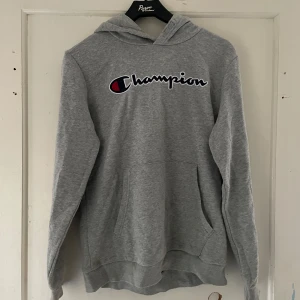 Grå hoodie från Champion - Säljer en grå hoodie från Champion. Den har en klassisk design med Champion-loggan på bröstet och en liten logga på ärmen. Tyvärr finns det några hål på baksidan, men den är fortfarande superbekväm och perfekt för chill dagar. Passar bra till både jeans och mjukisbyxor!