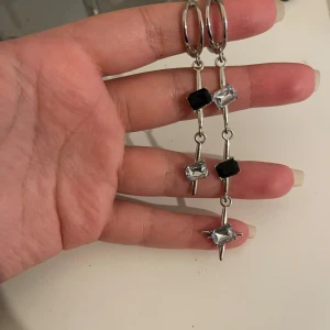 Silvriga örhängen med svarta och klara stenar - Säljer ett par snygga och unika silvriga örhängen(cuffs)med hängande svarta och klara stenar. De har en modern design med en liten ring upptill och flera stenar som hänger i rad. Perfekta för att ge en elegant touch till din outfit!