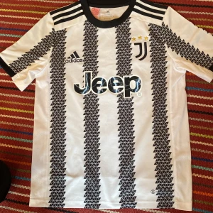 Juventus fotbollströja från Adidas - Säljer en snygg Juventus fotbollströja från Adidas. Den är vit med svarta detaljer och har det klassiska Juventus-emblemet på bröstet. Tröjan har korta ärmar och ett coolt mönster med svarta trianglar. Perfekt för alla Juventus-fans!