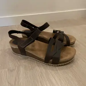 Säljer ett par snygga bruna sandaler med kilklack. De har en bekväm korksula och justerbara remmar för perfekt passform. Perfekta för sommardagar och ger en avslappnad men stilren look.