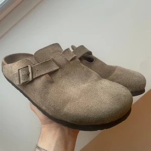Birkenstock Boston - Säljer ett par bekväma och stilrena Birkenstock Boston i beige mocka. De har en justerbar rem med spänne för perfekt passform. Perfekta för en avslappnad stil och passar bra till både vår och sommar.