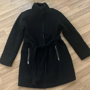 Svart kappa med bälte - Säljer en stilren svart kappa med bälte i midjan. Den har långa ärmar och dragkedjor på fickorna. Storklek M, aldrig andvänd 