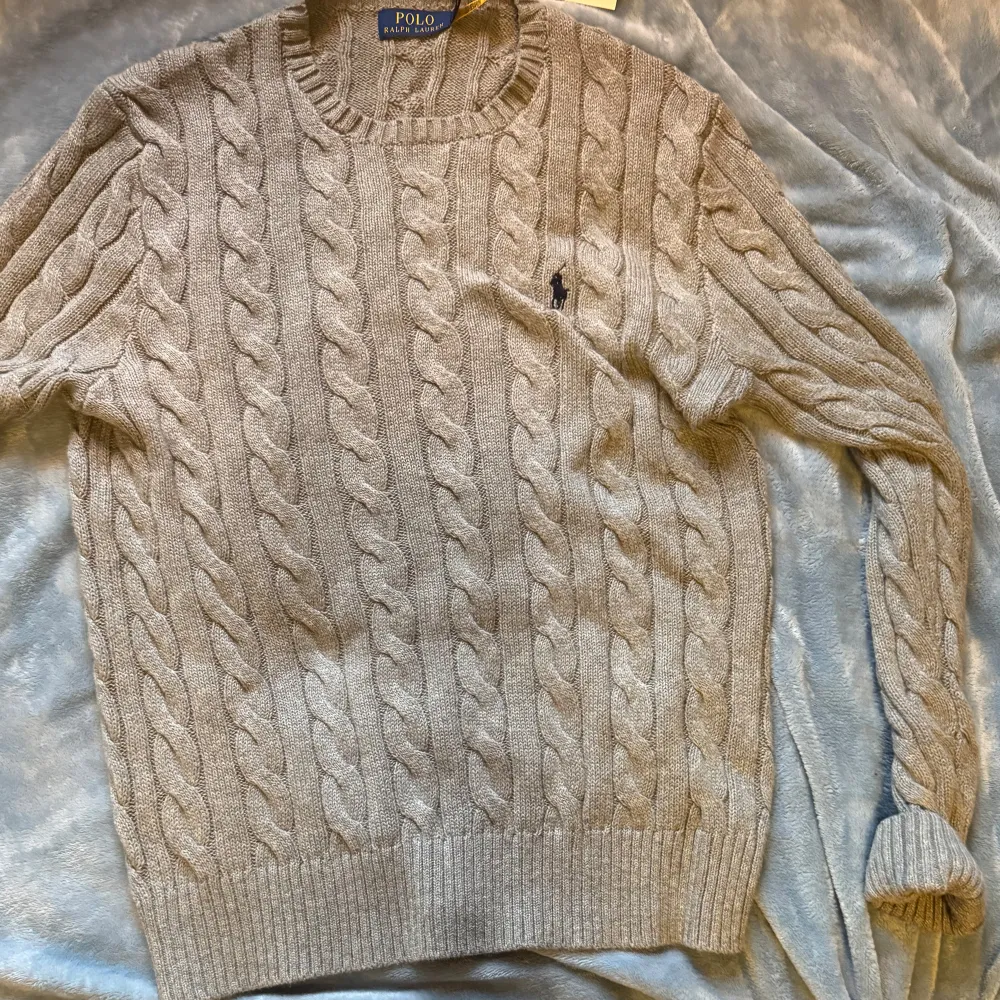 Säljer en superfin beige stickad tröja från Polo Ralph Lauren. Den har ett klassiskt flätmönster och är perfekt för både höst och vinter. Tröjan är långärmad och har en normal passform. Den är i nyskick och redo att hålla dig varm och stilren!. Tröjor & Koftor.