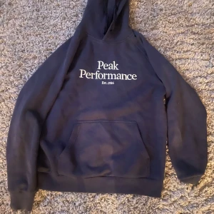 Blå hoodie från Peak Performance - Säljer en blå hoodie från Peak Performance med vit text på bröstet. Den har en klassisk känguruficka och en bekväm huva. Perfekt för en avslappnad stil.