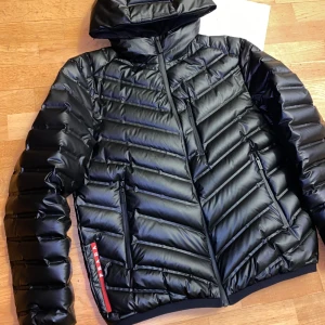 Svart dunjacka från Prada - Prada Nylon Light Hooded Puffer Jacket. Snygg svart dunjacka från Prada med huva och dragkedja. Jackan har en modern design med quiltade detaljer och är tillverkad i 100% polyamid. Perfekt för kyligare dagar. Nypris på Prada 30k.