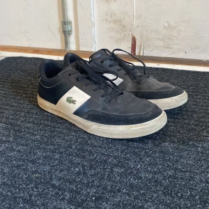 Svarta och vita sneakers från Lacoste - Snygga svarta och vita sneakers från Lacoste med klassisk krokodillogga på sidan. Skorna har snörning och en bekväm sula. Perfekta för en stilren look. Mycket använt, lite putts och rengöring skulle göra skon snyggare. (Hör av er angående pris) 