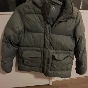 Grön pufferjacka från C - Säljer en grön pufferjacka från märket C. Jackan har en dragkedja och knappar framtill samt stora fickor. Den är långärmad och perfekt för kyligare väder. Insidan är fodrad med fleece för extra värme. Passar bra för både höst och vinter.