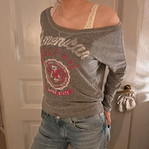 Grå offshoulder tröja från American Eagle - Säljer en grå offshoulder tröja från American Eagle med tryck i vitt och rött framtill. Tröjan har långa ärmar, finns ej att köpa i butik 