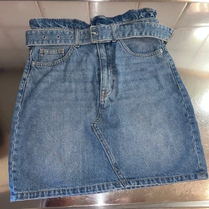 Jeanskjol med bälte från H&M - Snygg jeanskjol från H&M med medföljande bälte. Kjolen har en klassisk blå denimfärg och två bakfickor. Perfekt för en avslappnad stil.
