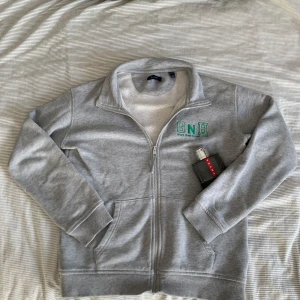Grå zip hoodie från Gant - Säljer en stilren grå zip hoodie från Gant med grönt broderi på bröstet. Tröjan har långa ärmar och en bekväm passform. Perfekt för en avslappnad look. Hör av dig vid intresse! Tröjan passar dig som är 155 till 162. Pris kan alltid diskuteras och fråga om du undrar något 