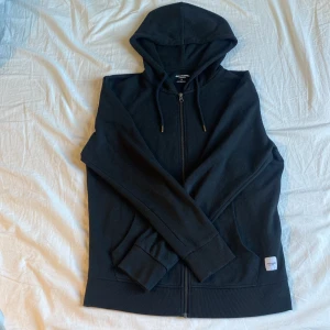 Svart hoodie från Jack & Jones - Säljer en svart hoodie från Jack & Jones Essentials. Den har en dragkedja framtill och justerbar huva med snören. Perfekt för en avslappnad look. Märkeslapp på framsidan ger en extra touch.