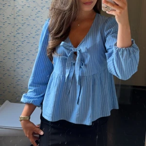 Blå randig blus med knytband - Säljer en söt blå blus från Vero Moda med vita ränder och knytband framtill. Blusen har långa ärmar och en v-ringad design som ger en avslappnad och stilren look. Perfekt för både vardag och speciella tillfällen.