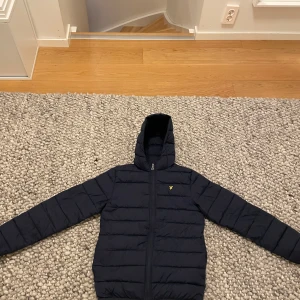 Mörkblå dunjacka från Lyle & Scott - Säljer en snygg mörkblå dunjacka från Lyle & Scott. Jackan har en dragkedja framtill och en praktisk huva. Perfekt vintern och vår jacka. En stilren jacka för ungdomar.