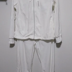 Vit mjukisbyxa och hoodie (pyjamas) från Colors by Venca - Säljer ett set med vita mjukisbyxor och matchande hoodie från Colors by Venca. Setet har en dragkedja framtill och snörning i midjan för justerbar passform. Perfekt för en avslappnad stil.