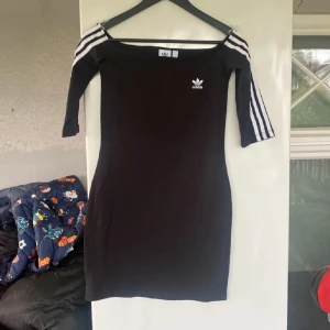 Svart klänning från Adidas - Snygg svart klänning från Adidas med trekvartsärm och de klassiska vita ränderna på ärmarna. Perfekt för en sportig och stilren look. Adidas-logga på bröstet ger en extra touch.