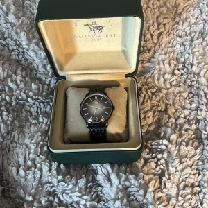 Svart klocka från Santa Barbara Polo & Racquet Club - Elegant svart klocka med mesh-armband och stilren urtavla. Klockan har tydliga siffror och datumvisning. Perfekt för en stilren look.