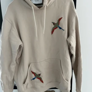 Beige hoodie från Axel Arigato - Säljer en stilren beige hoodie från Axel Arigato med broderade fåglar på bröstet och fickan. Tröjan har en klassisk känguruficka och justerbar huva med dragsko. Perfekt för en avslappnad stil.