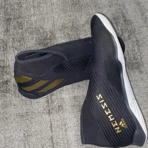 Adidas Nemeziz futsalskor  - Snygga svarta Adidas Nemeziz futsalskor med guldiga detaljer. Skorna har en strömlinjeformad design utan snörning och en slitstark vit sula. Perfekta för inomhusfotboll med sin greppvänliga yttersula.