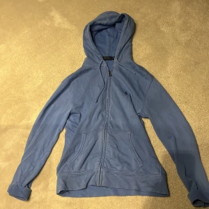 Blå hoodie från Ralph Lauren - Säljer en snygg blå hoodie från Ralph Lauren med dragkedja och broderad logga på bröstet. Köpt på NK för ca 2000kr använd fåtal gånger.