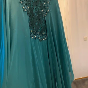 Turkos långklänning med spetsdetaljer - Elegant turkos långklänning med vackra spetsdetaljer och pärlor på bröstet och ärmarna. Klänningen har en draperad design och långa ärmar, vilket ger en flytande och sofistikerad look. Perfekt för speciella tillfällen.