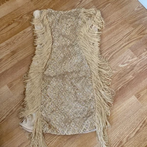 Guldig klänning med fransar från PrettyLittleThing - Snygg guldig klänning från PrettyLittleThing med glittrigt mönster och fransar längs sidorna. Axelbandslös design som ger en elegant look. Perfekt för att sticka ut på festen! ✨