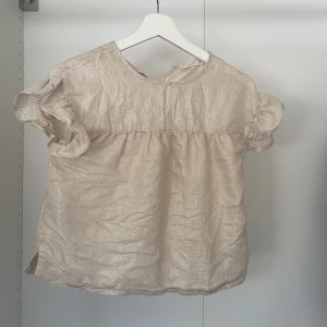 Beige glittrig blus från Mango Kids - Säljer en söt beige blus från Mango Kids med glittriga ränder. Blusen har korta ärmar med volangdetaljer och en knapp i nacken. Perfekt för en festlig look! ✨