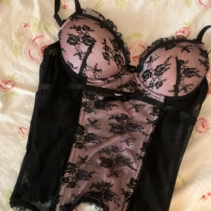 Svart och rosa korsett från Ann Christ -  svart och rosa korsett från Ann Christ med blommigt spetsmönster. Den har justerbara axelband och en dekorativ rosett framtill.