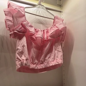 Rosa volangtopp - Säljer en söt rosa topp med volangdetaljer. Toppen är kortärmad och har en luftig passform, perfekt för varma dagar. Den är gjord i ett lätt och bekvämt material.