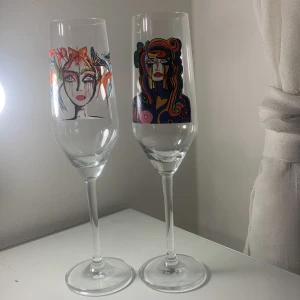 Champagneglas med konstnärligt motiv - Två unika champagneglas med färgglada konstnärliga motiv av ansikten. Det ena glaset har ett motiv med varma färger som rött och orange, medan det andra har ett motiv med en mix av färger som blått, rosa och grönt. Perfekta för att ge en konstnärlig touch till din dukning.