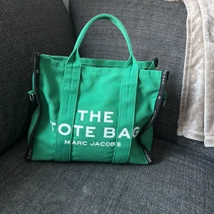Grön tote bag från Marc Jacobs - Snygg grön tote bag från Marc Jacobs med texten 'THE TOTE BAG' på framsidan. Väskan har praktiska handtag och en dragkedja upptill. Perfekt för att bära med sig allt man behöver under dagen.