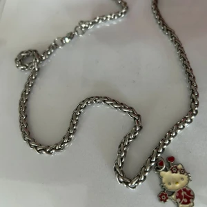 Silverfärgat halsband •Hello Kitty - Charmigt silverfärgat halsband med en söt kattberlock. Berlocken har röda detaljer och små blommor, vilket ger en lekfull och unik touch. Perfekt för kattälskare eller som en rolig accessoar till din outfit.