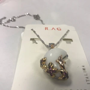 Snygg Halsband med vit och guldberlock  - Elegant halsband från R.A.G med en vit berlock dekorerad med guldfärgade detaljer och små lila stenar. Kedjan är silverfärgad och har en fin glans. Perfekt för att ge en touch av lyx till din outfit.  Helt ny 