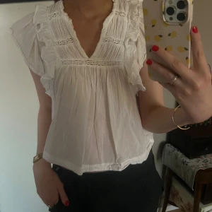 Vit blus med volangdetaljer - Charmig vit blus med volangdetaljer och v-ringning. Blusen har korta ärmar och en luftig passform, perfekt för varmare dagar. Den gulliga stilen gör den lätt att matcha med olika outfits.