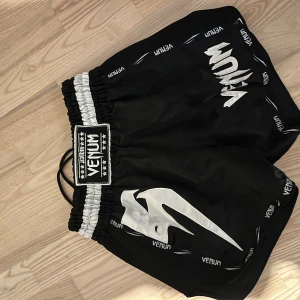 Venum thaishorts - Säljer ett par svarta träningsshorts från Venum med vit logga och elastisk midja. Perfekta för intensiva träningspass. Shortsen har en sportig look med vita detaljer och är gjorda för att ge maximal rörelsefrihet.