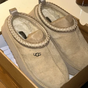 Beige mockaskor från UGG - Säljer ett par bekväma beige mockaskor från UGG med mjukt foder inuti. Skorna har en stilren design med UGG-loggan på sidan och en dekorativ kant runt öppningen. Ugg tazz modellen, de är i bra skick 