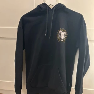 Svart hoodie från Yourturn med tryck - Säljer en svart hoodie från Yourturn med ett coolt tryck av en vädursskalle på ryggen och ett mindre tryck på bröstet. Tröjan har en klassisk känguruficka och justerbar huva med snören. Perfekt för en avslappnad stil.