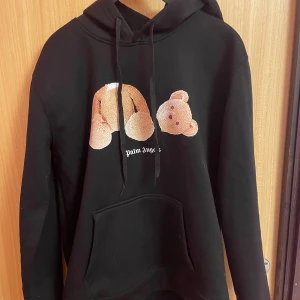 Svart hoodie från Palm Angels - Säljer en svart hoodie från Palm Angels med en unik nallebjörnsdesign på framsidan. Hoodien har en stor ficka framtill och texten 'Palm Angels' tryckt på både fram- och baksidan. Perfekt för en avslappnad stil.