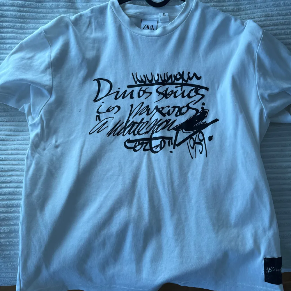 Säljer en vit t-shirt från Zara med ett coolt svart grafiskt tryck på framsidan. Perfekt för en avslappnad stil. T-shirten har en klassisk rund hals och korta ärmar.. T-paidat.