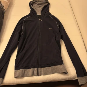 Mörkblå huvtröja från Boss - Säljer en stilren mörkblå huvtröja från Boss med dragkedja och grå detaljer. Tröjan har en bekväm passform och är perfekt för kyligare dagar. Den har långa ärmar och en praktisk huva.