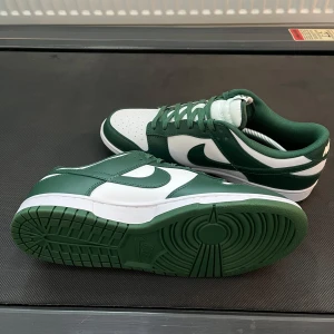 Nike Dunk i grönt och vitt - Snygga Nike Dunk sneakers i en fräsch grön och vit färgkombination. Skorna har klassisk snörning och en bekväm passform. Perfekta för en stilren look.